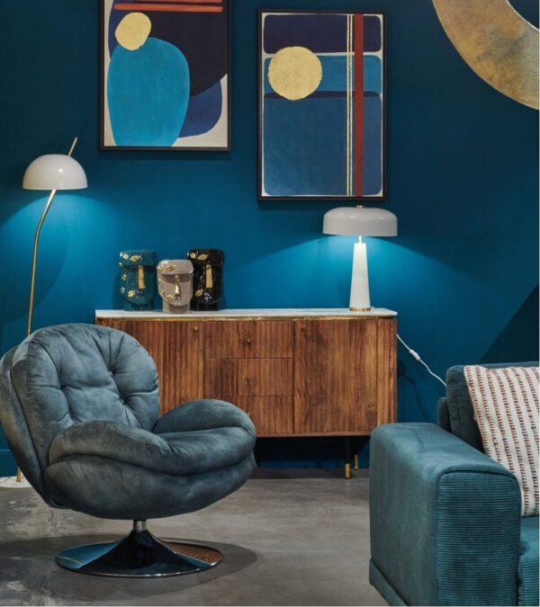 Fauteuil Memento en velours effet vieilli couleur pétrole, à assise enveloppante et base pivotante chromée, mis en valeur dans un salon contemporain aux tons bleus profonds et mobilier en bois rétro.
